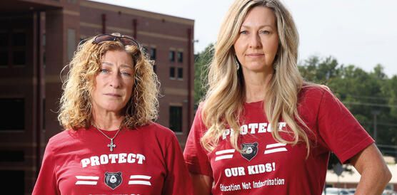 Zwei mittelalte Frauenstehen nebeneinander beide schulterlanges Haar und das gleiche T-Shirt Protect our kids steht auf denen